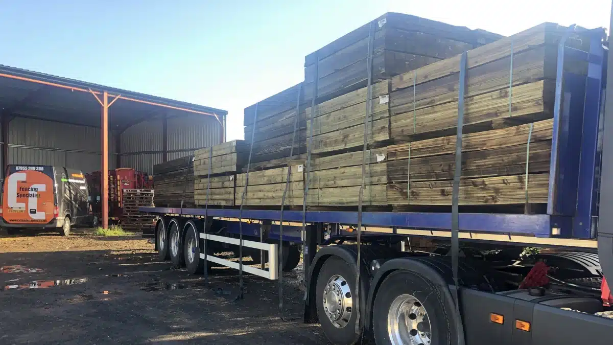 Flatbed lorry loaded with stacked timber in a yard, with a fencing specialist van nearby