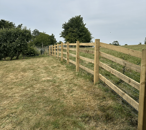 Wooden post-and-rail fence running along a grassy field with trees in the background