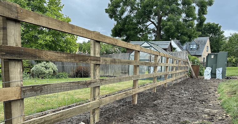 New wooden fence along a muddy garden boundary, with houses and a greenhouse behind it