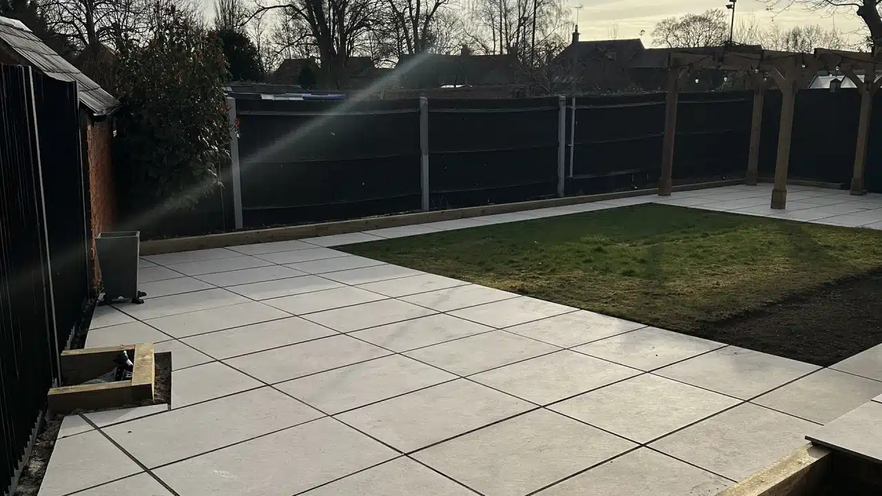 Patio with large grey paving slabs around a small lawn, bordered by a dark fence and pergola