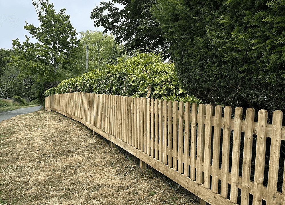 Wooden picket fence along a roadside verge with trees and shrubs behind it