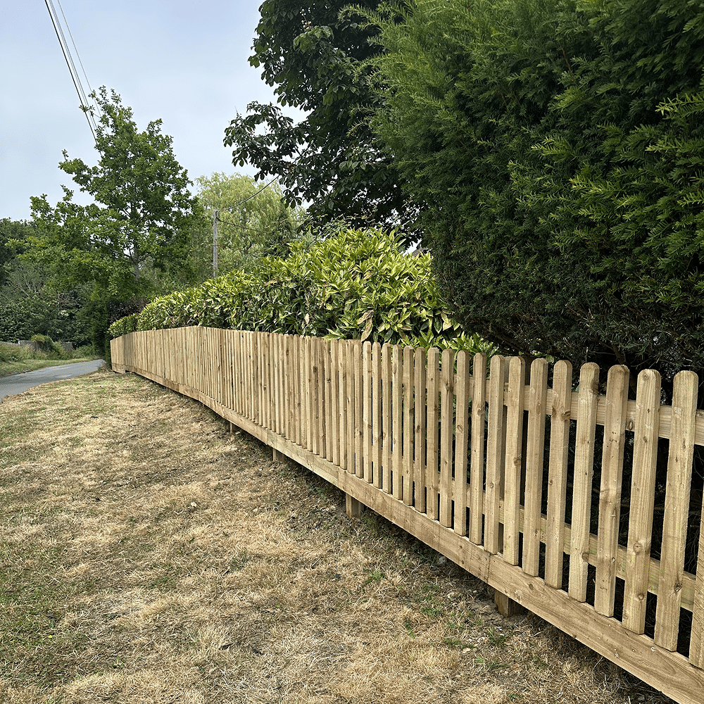 Wooden picket fence along a roadside verge with trees and shrubs behind it