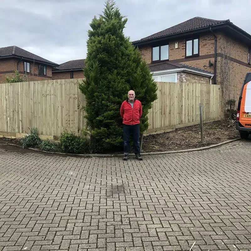 Man standing in front of a large evergreen tree beside a suburban house and fence