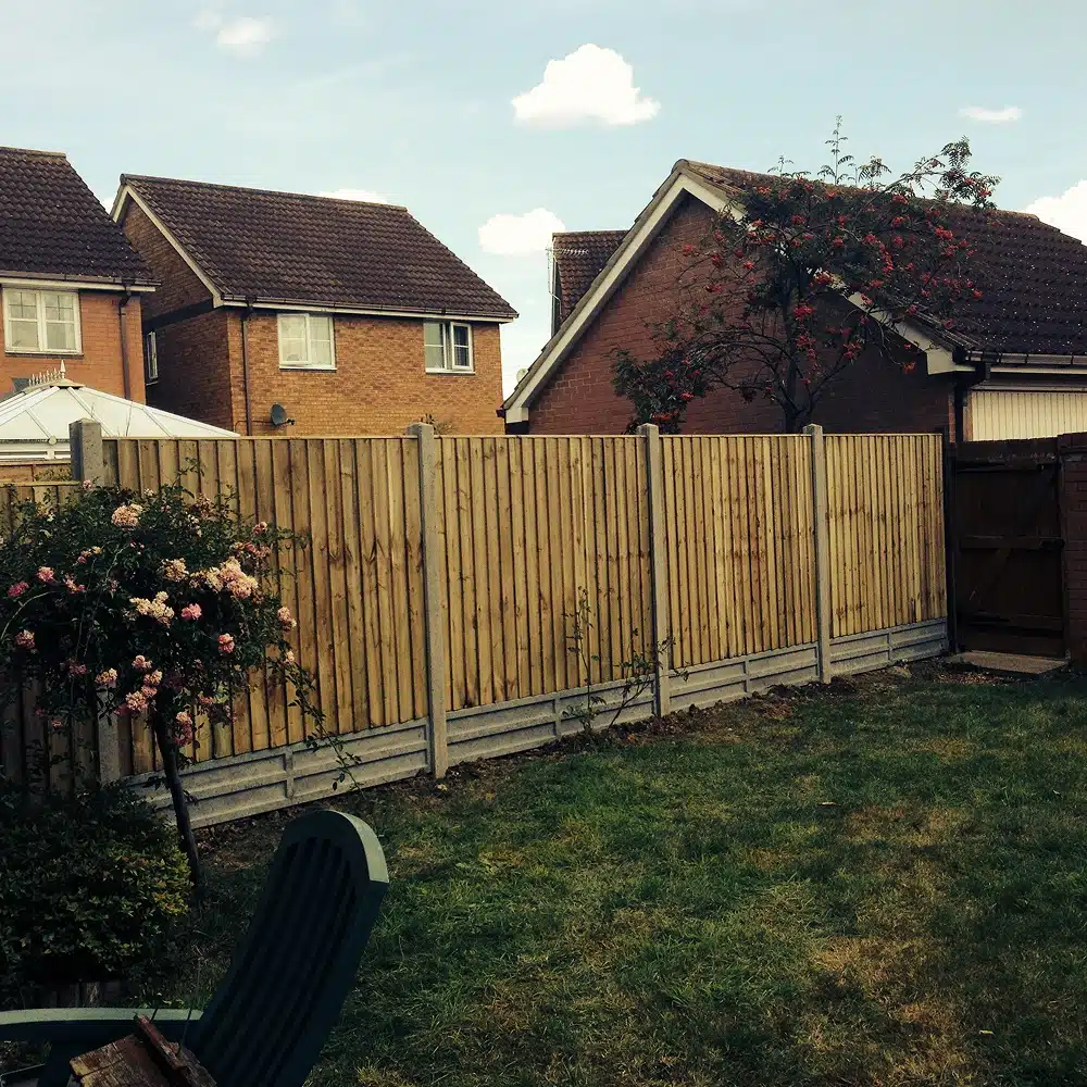 Back garden with a wooden fence, lawn, flowering shrubs and neighbouring houses behind it