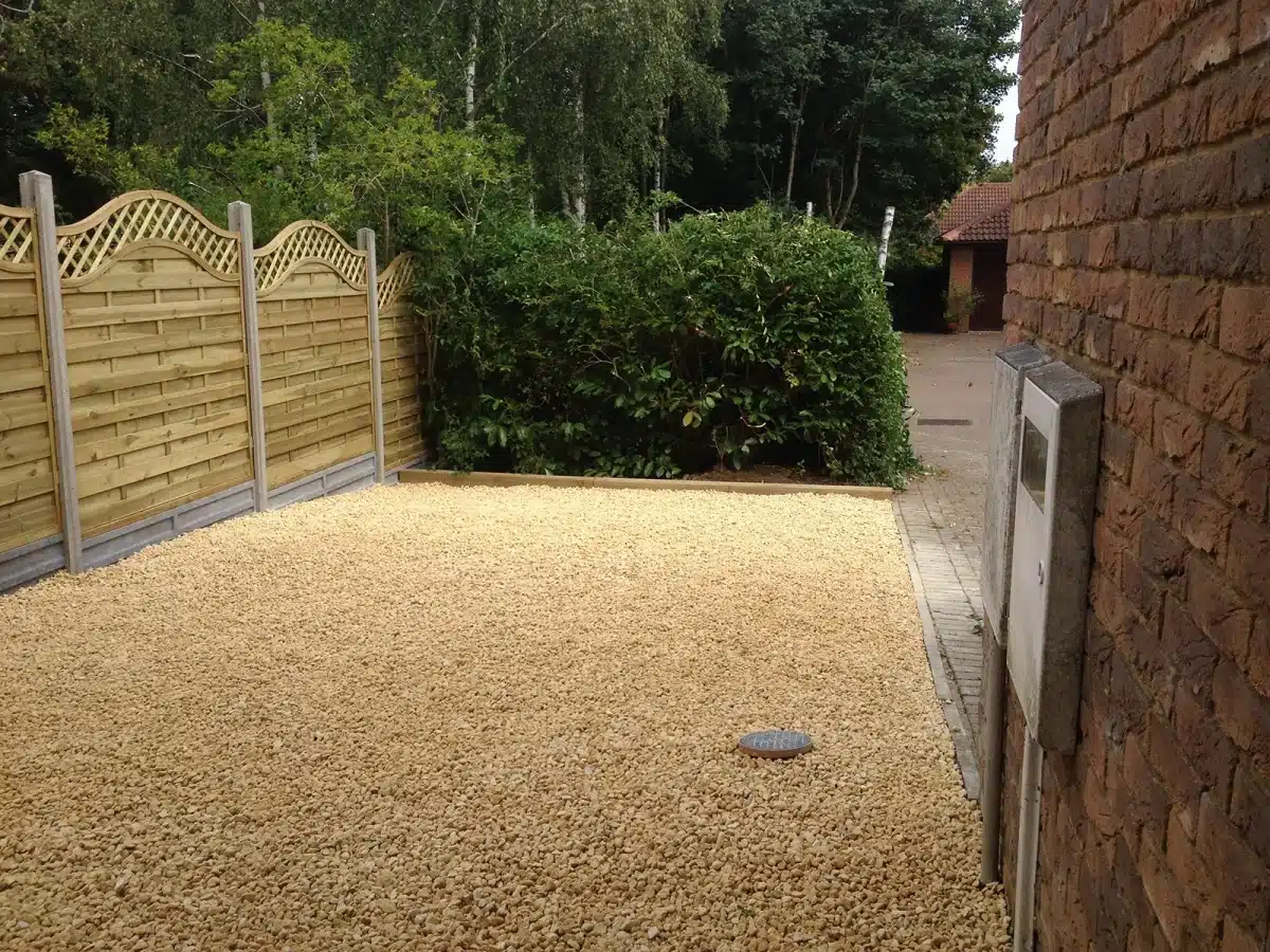 Gravel-covered driveway beside a brick house, with a wooden fence and hedge