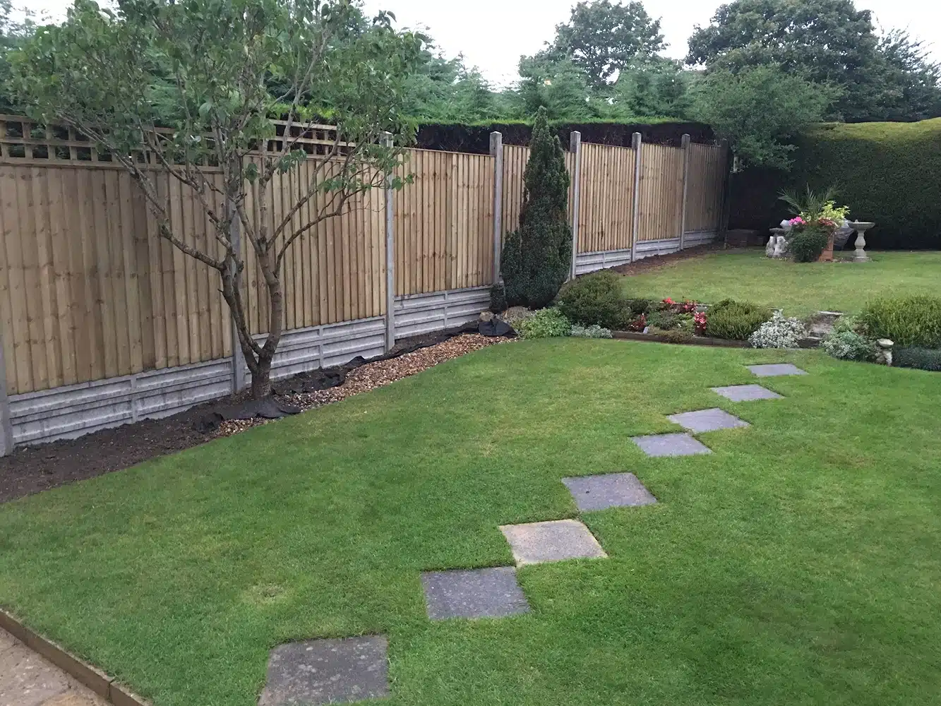 Back garden with lawn, stepping stones, wooden fence and planted borders
