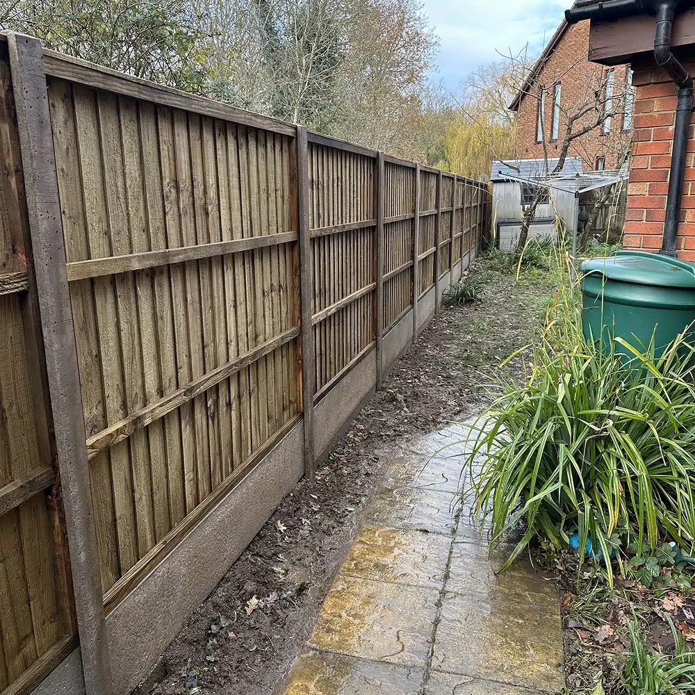 Narrow muddy side passage beside a wooden fence and brick house, with a wet paved path and garden plants