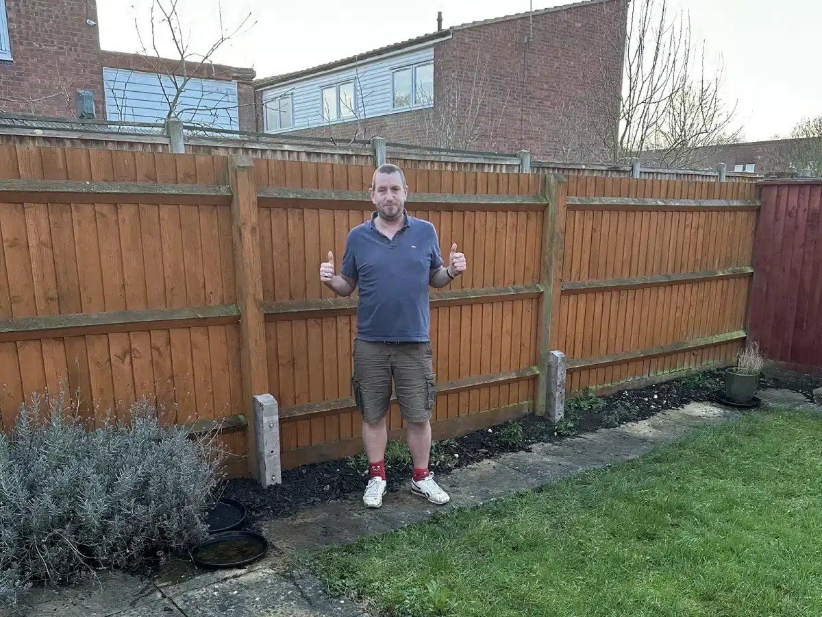Man standing in a garden giving two thumbs up in front of a wooden fence
