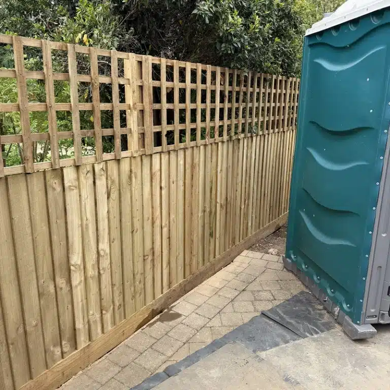 Wooden fence beside a portable toilet on a paved path