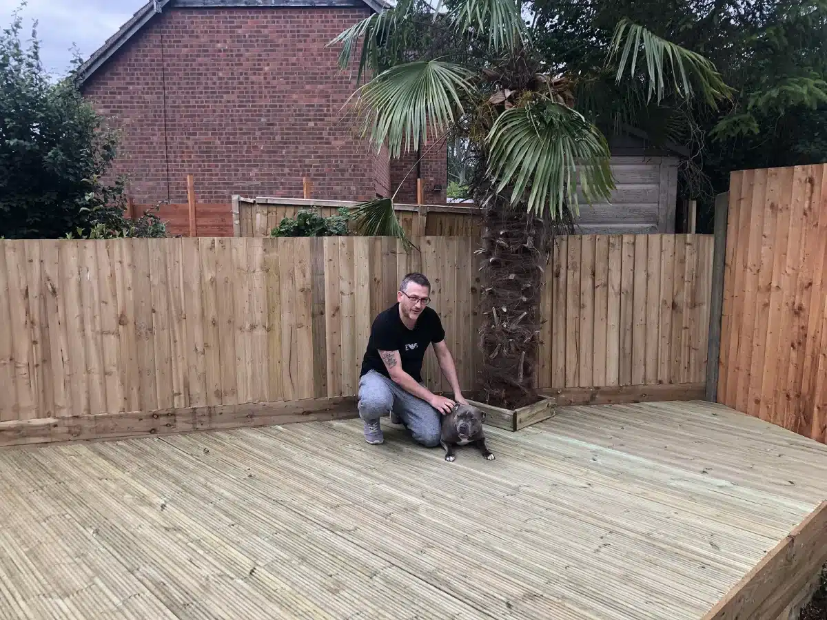 Man kneeling beside a dog on a newly built wooden deck in a fenced garden