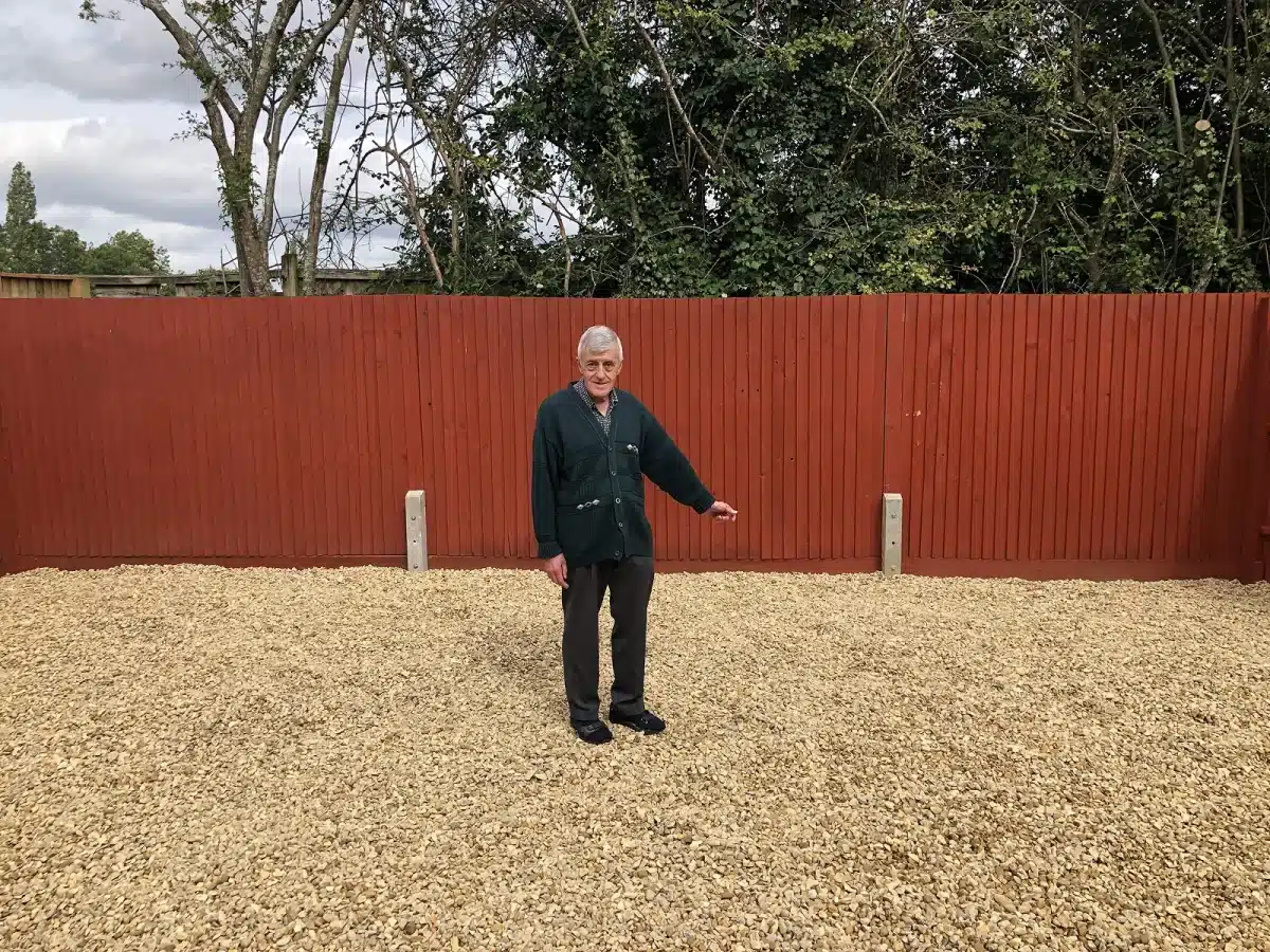 Man standing on a gravel surface in front of a red wooden fence and trees